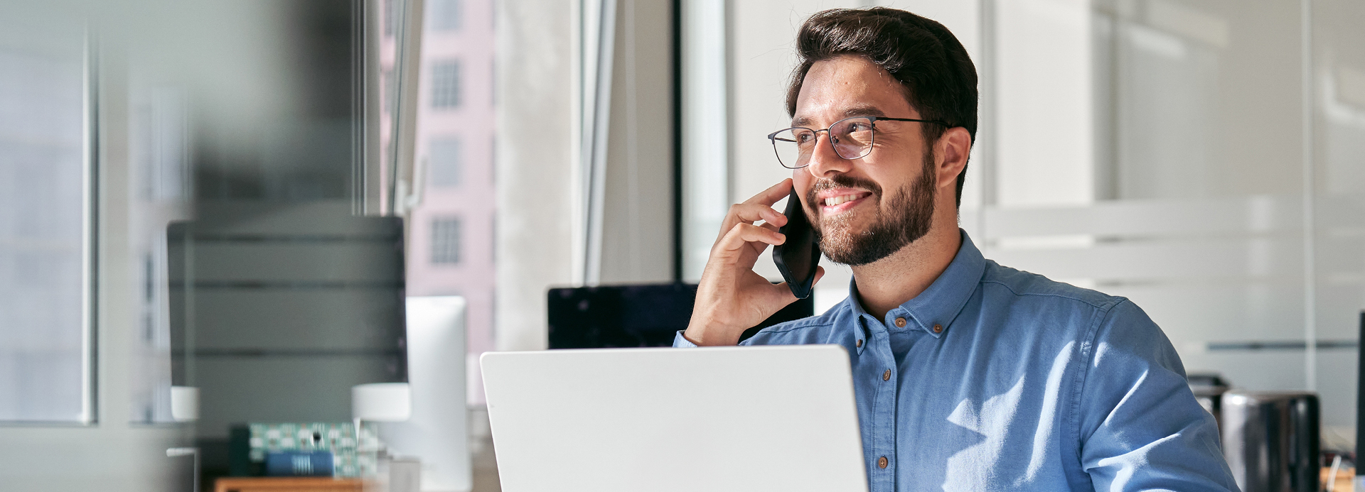 Mann mit Brille und blauem Hemd sitzt an einem Schreibtisch und telefoniert mit einem Smartphone