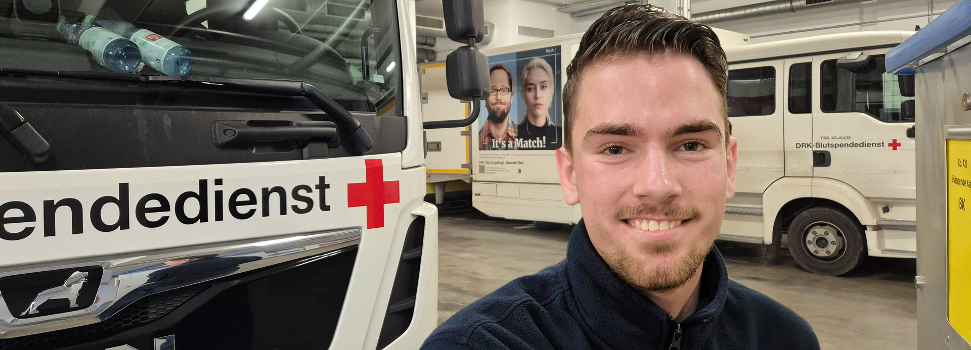 Junger Mann mit dunkelblauer Jacke vor zwei LKWs in der Lagerhalle