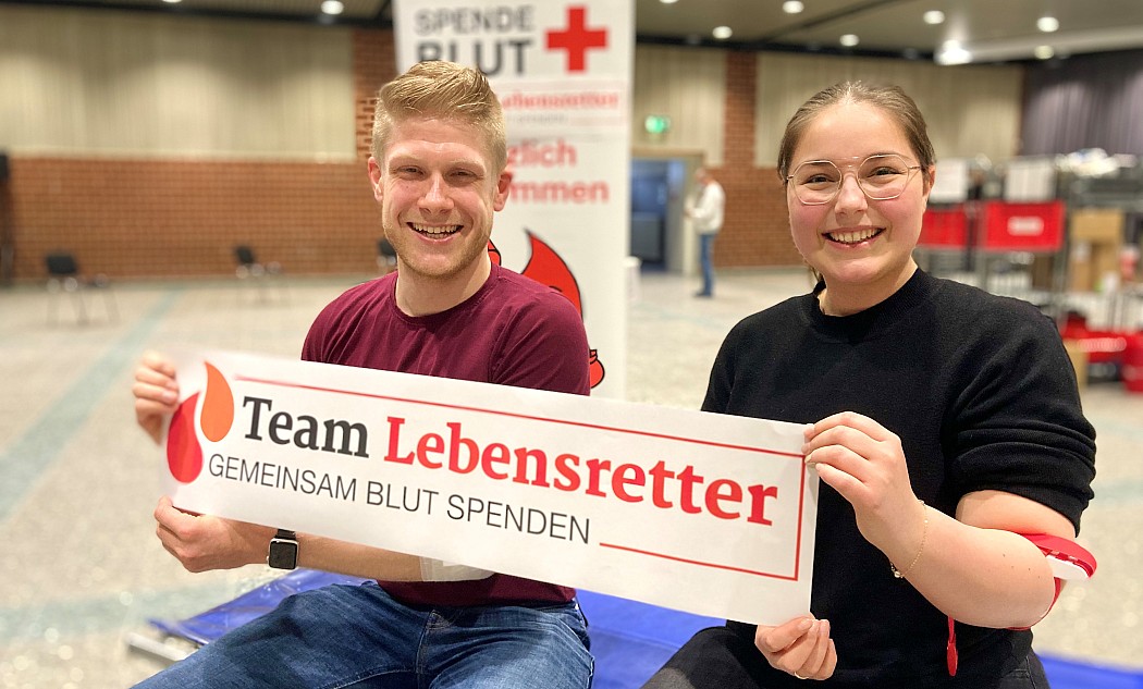 Zwei Personen Blutspender sitzen auf einer Liege und halten Plakat Logo Team Lebensretter Gemeinsam Blut spenden