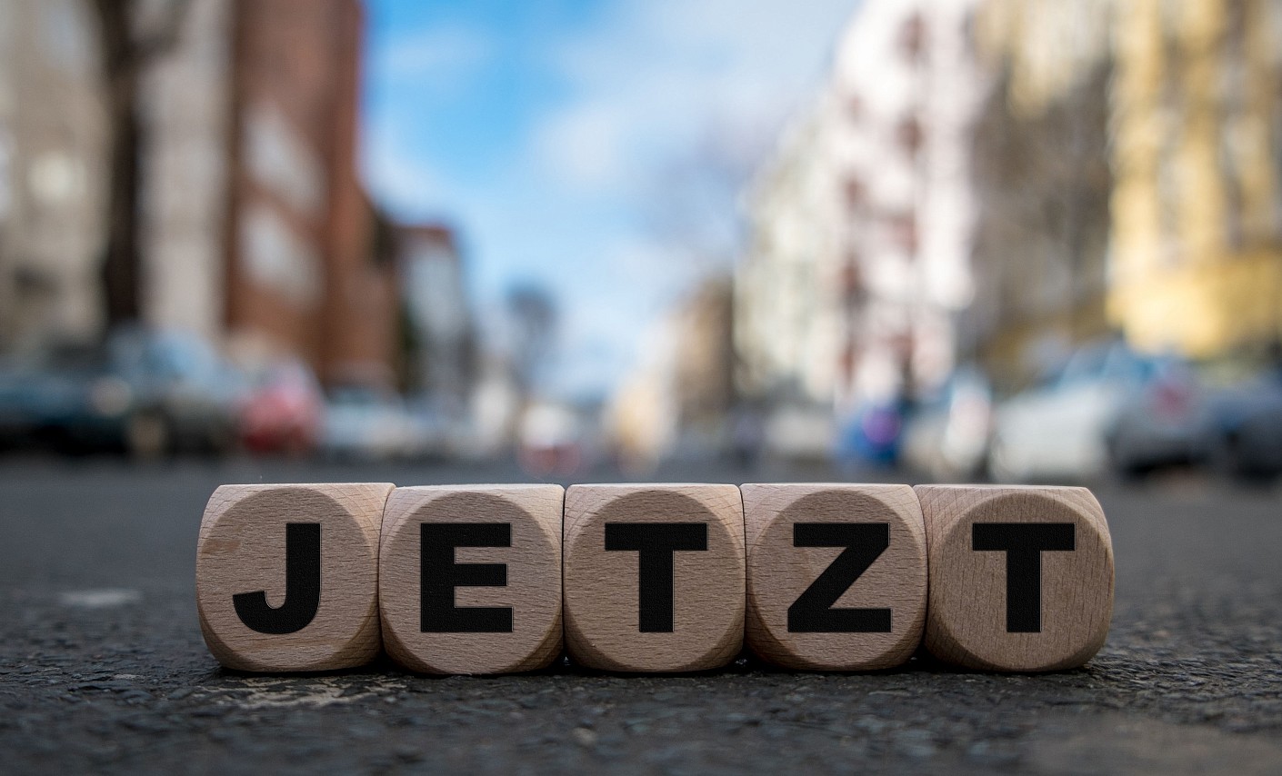 "Jetzt" als Buchstrabenblock vor einer Straßenszene