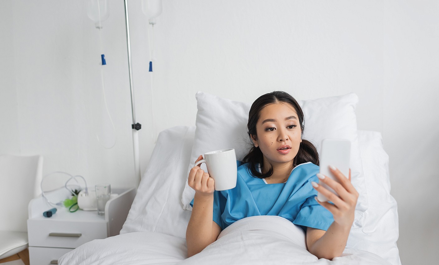 Patientin sitzt mit Handy und Kaffeetasse im Bett