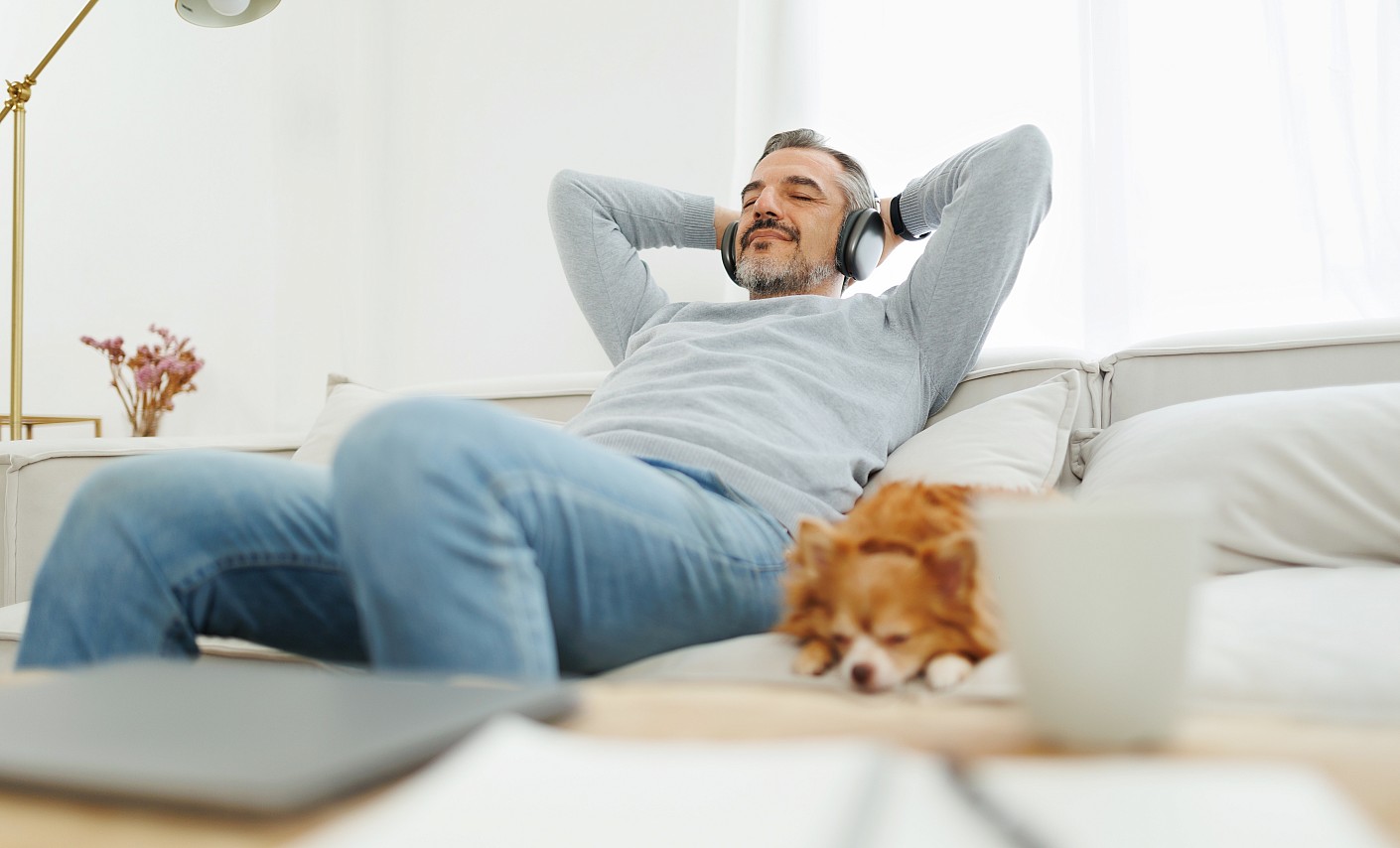 Ein Mann mittleren Alters sitzt mit Hund auf dem Sofa und hört Musik über Kopfhörer Ein Mann mittleren Alters sitzt mit Hund auf dem Sofa und hört Musik über Kopfhörer
