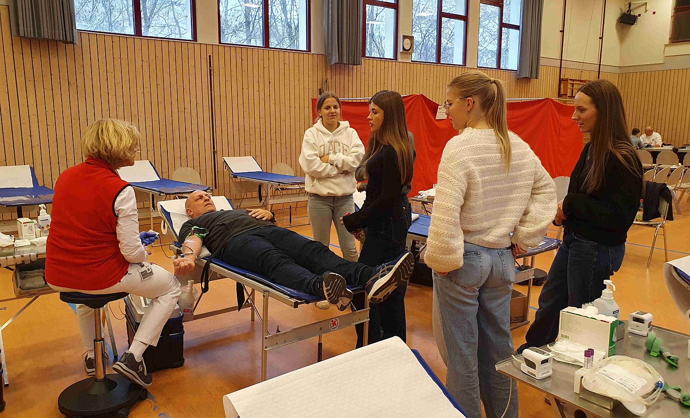 Entnahmeschwester erklärt den Schülerinnen den Ablauf der Blutspende von Dominick Thiel, der gerade Blut spendet