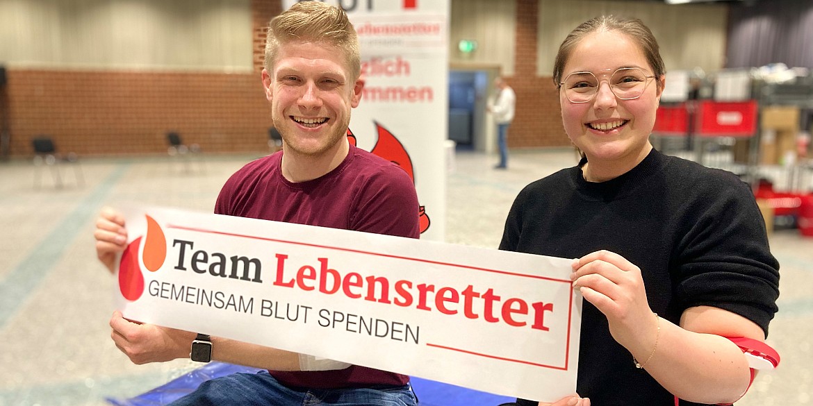 Zwei Personen Blutspender sitzen auf einer Liege und halten Plakat Logo Team Lebensretter Gemeinsam Blut spenden