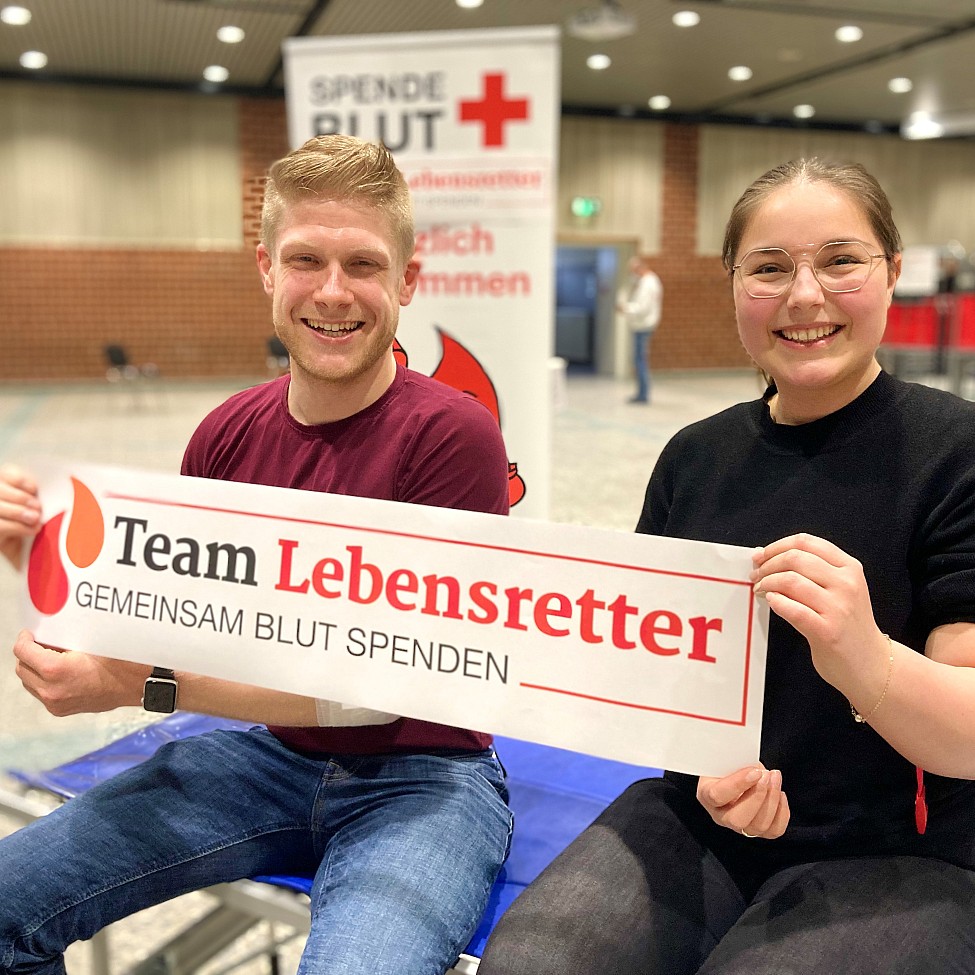 Zwei Personen Blutspender sitzen auf einer Liege und halten Plakat Logo Team Lebensretter Gemeinsam Blut spenden